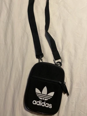 Svart axelväska från Adidas - Svart axelväska från Adidas med klassiska treklöver-loggan i vitt framtill. Väskan har justerbar axelrem, dragkedja och ett extra fack på framsidan. Tillverkad i slitstark polyester och har detaljer i svart plast. Perfekt storlek för mobil, plånbok och nycklar.