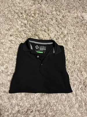 Svart piké från Oscar Jacobson - Klassisk svart långarmad pikétröja från Oscar Jacobson med krage och knappar framtill. Snygg och enkel design med diskreta detaljer i grått på insidan av kragen. Perfekt till jeans eller shorts.