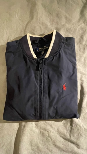 Marinblå bomberjacka från Polo Ralph Lauren - Säljer en marinblå bomberjacka från Polo Ralph Lauren. Storlek 14-16. Två små slitmärken vid ärmarna. 