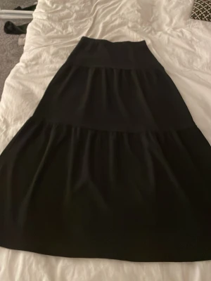 Svart långkjol från Kappahl - En svart långkjol från Kappahl med bred midja och volangdetaljer. Kjolen har en enkel och stilren look som passar till många olika outfits. Tillverkad i ett mjukt och lätt material som ger en skön känsla.