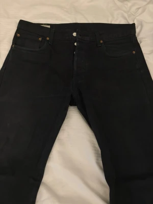 Levi's 501 svarta jeans - Säljer ett par klassiska svarta Levi's 501 jeans med raka ben och knappgylf. Jeansen har fem fickor, silverfärgade knappar och den ikoniska röda Levi's-lappen bak. Perfekta för dig som gillar tidlös stil och vill ha ett par jeans som funkar till allt. Storlek 34/32