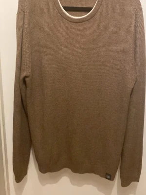 Brun stickad tröja med rund hals - En enkel och stilren brun stickad tröja med rund halsringning. Tröjan har lång ärm och en diskret etikett nertill. Perfekt för lager på lager och passar till både jeans och chinos.