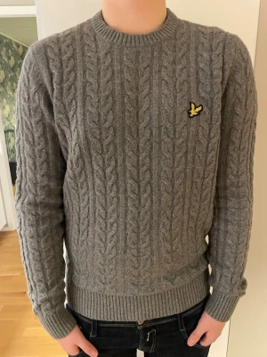 Grå kabelstickad tröja Lyle & Scott - Säljer en grå kabelstickad tröja från Lyle & Scott med rund hals och den klassiska gula loggan på bröstet. Tröjan har ribbade muddar och nederkant, lång ärm och är gjord i mjukt material som passar perfekt till höst och vinter.