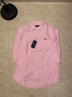 Rosa skjorta från Ralph Lauren - Klassisk rosa skjorta från Ralph Lauren med button-down krage och broderad logga på bröstet. Skjortan har långa ärmar, vita knappar och är tillverkad i mjuk bomull. Perfekt för dig som vill ha en clean och stilren look. Priset kan diskuteras 😊