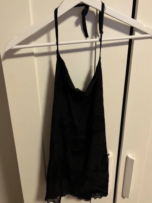 Svart drakmönstrad linnetopp - Säljer en svart linnetopp med tunna axelband som knyts i nacken. Toppen har ett snyggt drakmönster i sammet och mesh-liknande material samt spetskant längst ner. 