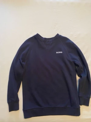Mörkblå sweatshirt från Björn Borg - Säljer en stilren mörkblå sweatshirt från Björn Borg med enkel design och liten vit logga på bröstet. Tröjan har rund halsringning, långa ärmar och breda muddar vid ärmslut och nederkant. Perfekt för en chill och sportig look.