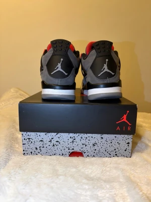 Nike Air Jordan 4 Retro grå/röd - Nike Air Jordan 4 Retro sneakers i grå mocka med svarta och röda detaljer. Skorna har en vit och svart platt sula, meshpaneler på sidorna och klassisk Jumpman-logga på hälen. Röd insida och snörning framtill ger en fet kontrast. Köpt för 4499kr.