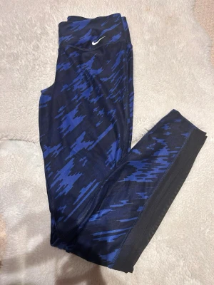 Nike Dri-Fit blå camo leggings - Nike Dri-Fit leggings i blått och svart camo-mönster. Snyggt tight passform med mesh-detaljer på benen för extra ventilation. Hög midja och stretchigt syntetmaterial som är perfekt för träning. Ikonisk Nike-logga framtill.