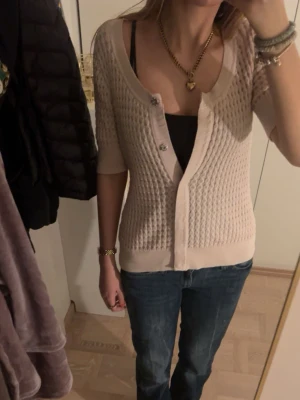 Beige stickad kofta med kort ärm - Säljer en beige stickad kofta med kort ärm och snyggt mönster. Koftan har u-ringning, knappar framtill och ribbade muddar.