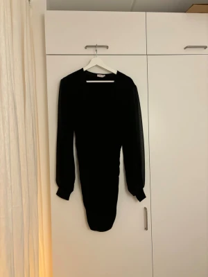 Svart omlottklänning med lång ärm - Säljer en svart kortklänning från NA-KD med omlott-design och v-ringad hals. Klänningen har långa, lätt transparenta ärmar med breda muddar och en figurnära passform. Perfekt för dig som gillar stilrena och trendiga plagg.