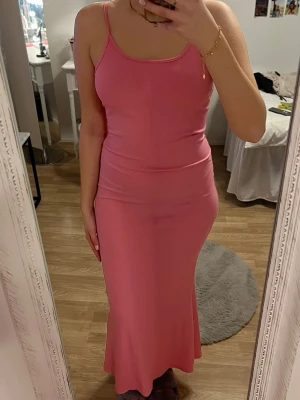 Rosa ribbad långklänning - Säljer en snygg rosa långklänning med smala axelband och ribbad struktur. Klänningen har en figurnära passform och är tillverkad i ett mjukt, stretchigt material som sitter skönt på kroppen. Storlek XS men passar S/M med. Jag är 170cm lång och klänningen går ner till mina hälar.