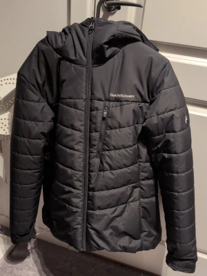 Svart Peak Performance pufferjacka XL - Svart pufferjacka från Peak Performance i storlek XL. Jackan har huva, quiltad design och dragkedja framtill samt en sned bröstficka med dragkedja. Perfekt för kalla dagar och har en clean, sportig look. Använd 1 gång.