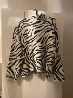 Zebramönstrad stickad polotröja HM  - Säljer en fluffig stickad polotröja med zebramönster i svart och vitt. Tröjan har en hög krage och långa ärmar, perfekt för dig som vill sticka ut med en trendig och cozy look. SLUTSÅLD 