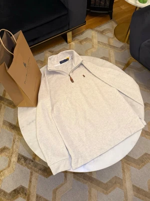 Ljusgrå half zip från Polo Ralph Lauren - Snygg ljusgrå långärmad tröja från Polo Ralph Lauren med half zip och klassisk broderad logga på bröstet. Tröjan har hög krage och är tillverkad i mjuk bomull, perfekt för lager-på-lager under höst och vinter.