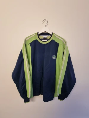 Vintage adidas 90 tal tjocktröja - Retro Adidas 90 tals sweatshirt i marinblått och ljusgrönt med vita detaljer längs ärmarna. Klassisk rund halsringning och Adidas-logga på bröstet. Oversized passform och långa ärmar, perfekt för en sportig och avslappnad stil.