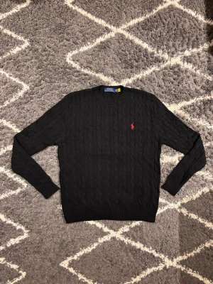 Polo Ralph Lauren tröja svart  - Svart kabelstickad tröja från Polo Ralph Lauren med klassisk rund hals och röd broderad logga på bröstet. Tröjan har långa ärmar och ribbade muddar vid ärmslut och nederkant. Jag väger 70KG och är 180CM och den passar mig perfekt.