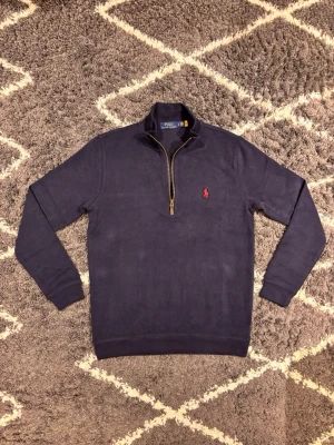 Polo Ralph Lauren half zip - Säljer en marinblå långärmad tröja från Polo Ralph Lauren med klassisk röd logga på bröstet. Tröjan har en snygg half zip-dragkedja och ribbade muddar. Jag väger 70KG och är 180CM och den passar mig perfekt.