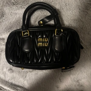 Svart quiltad handväska - Snygg svart handväska i quiltat skinnliknande material med guldiga detaljer och logga framtill. Väskan har dubbla handtag, justerbar axelrem och dragkedja upptill. Perfekt storlek för det viktigaste och ger en lyxig känsla till din outfit.