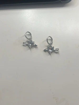 Silvriga örhängen med hjärta och pil - Säljer ett par silvriga örhängen med hjärtformad sten och pil-detalj. Örhängena har en cool och edgy vibe, perfekta för att lyfta vilken outfit som helst. Hängs på örat med enkel ring och har en glansig finish.