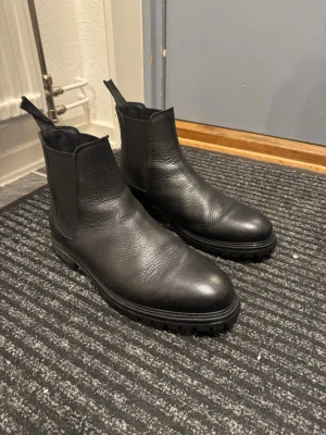 Matinicue  - Säljer ett par klassiska svarta chelsea boots i skinn med elastiska paneler på sidorna och dragflikar både fram och bak. De har en rund tå och grov sula som ger ett stilrent men robust intryck. Perfekta till höstens och vinterns outfits.