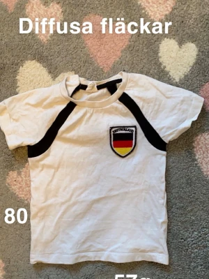 Vit t-shirt med Tyskland-emblem - Vit t-shirt med svarta detaljer och broderat Tyskland-emblem på bröstet. Klassisk passform och korta ärmar, perfekt för dig som gillar sportig stil. 🌸 Diffusa fläckar . Storlek 80