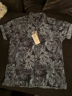 Blå mönstrad skjorta Jack & Jones - Säljer en kortärmad skjorta från Jack & Jones i mörkblått med ljusblått bladmönster. Skjortan har avslappnad passform, klassisk krage och knappar framtill. Perfekt för dig som gillar en chill och snygg stil. Helt ny med prislappen kvar! Nypris 499kr