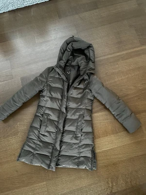 Grå dunjacka med huva från Zara Basic - Snygg grå dunjacka från Zara Basic i storlek M. Jackan är quiltad, har huva och dragkedja framtill. Lång modell med raka linjer och praktiska fickor. Perfekt för kalla dagar och enkel att matcha med olika outfits.