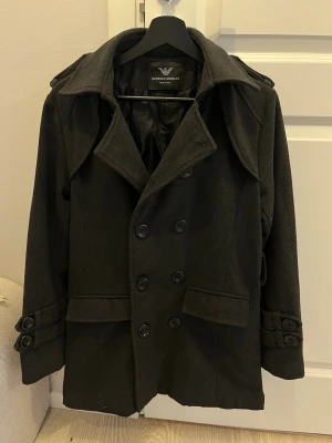 Svart rock från Giorgio Armani - Stilren svart rock från Giorgio Armani med dubbelknäppning, bred krage och axelklaffar. Jackan har snygga detaljer med knappar vid ärmslut och fickor framtill. Tillverkad i ullblandning och fodrad för extra komfort. Perfekt för dig som vill ha en klassisk och tidlös look.