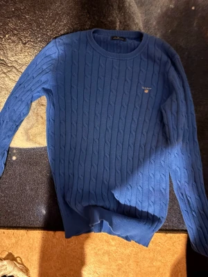Blå tröja från Gant - Säljer en blå kabelstickad tröja från Gant med rund halsringning och diskret logga broderad på bröstet. Tröjan har lång ärm och ribbade muddar vid ärmslut och nederkant. Perfekt för dig som gillar klassisk och stilren look.