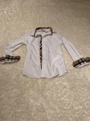 Vit Burberry skjorta med rutiga detaljer - Säljer en klassisk vit skjorta från Burberry med ikoniska beige, bruna och röda rutiga detaljer på krage, knappslå och manschetter. Skjortan har lång ärm, normal passform och knappar framtill. Perfekt för dig som vill ha en clean look med lyxig touch.