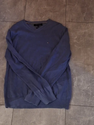 Blå tröja från Tommy Hilfiger - Mörkblå långärmad tröja från Tommy Hilfiger med v-ringning och diskret logga på bröstet. Tröjan är i mjuk bomull och har ribbade muddar vid ärmslut och nederkant. Perfekt för lager-på-lager under kyliga dagar.