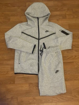 Nike tech set  - Säljer ett par grå mjukisbyxor från Nike med svart logga på benet och dragsko i midjan. Byxorna är i mjukt material och har en avslappnad passform, perfekta för chill eller träning. Matchar snyggt med hoodie i samma färg. Funkar i både storlek S och storlek M