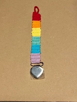 Virkad regnbågsfärgad napphållare - Handgjord napphållare i virkad bomull med regnbågsfärger: röd, orange, gul, grön, blå och lila. Metallklämma i form av ett hjärta längst ner. Perfekt accessoar för att hålla koll på nappen.