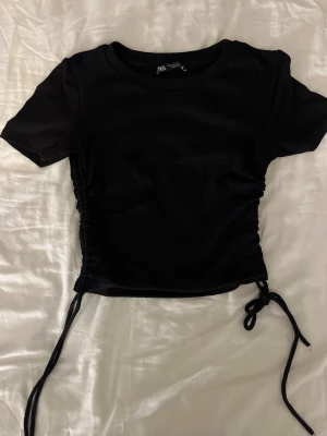Svart topp med scrunch från Zara - Säljer en svart topp med snörning på sidorna från Zara. Toppen har en rund sopringad hals och är tillverkad i ett mjukt, ribbat material som sitter snyggt på kroppen. 