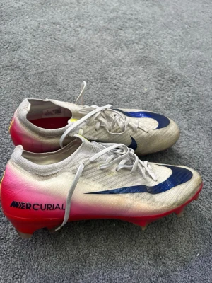Nike Mercurial fotbollsskor strl 42,5 - Nike Mercurial fotbollsskor i vitt med röd swoosh och rosa/röd sula. Skorna har snörning och är tillverkade i syntetmaterial för lätt känsla på planen. Perfekta för dig som vill ha snabbhet och kontroll på fotbollsplanen. Köpta för 3199 kr, använda och skicket är 7,5/10. Därför jag säljer för billigare pris! Se defekt på sista bilden.