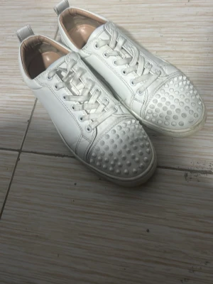 Vita sneakers med nitar från Louboutin - Säljer ett par vita sneakers från Louboutin i skinn med ikoniska röda sulor och coola nitar på tån. Skorna har snörning och en klassisk rund tå. Perfekta för dig som vill sticka ut med en lyxig och edgy vibe. En spik saknas men syns inte eftersom jag har målat över☺️