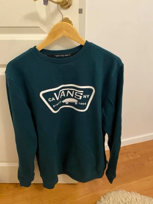 Mörkgrön crewneck från Vans - Säljer en mörkgrön långärmad crewneck från Vans med stor vit logga framtill. Tröjan har rund halsringning och är gjord i mjuk bomull, perfekt för chill dagar. Klassisk streetwear-stil med enkel design och skön passform.