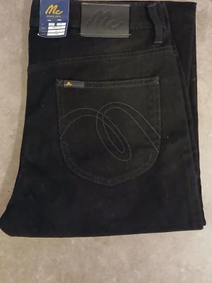 Svarta jeansbyxor från Mc, W34 L31 - Svarta jeansbyxor från Mc med klassisk femficksdesign och snygg broderad bakficka. Raka ben och normal passform. Materialet är slitstarkt jeans och byxorna har en stilren look med diskreta detaljer. Perfekt för dig som gillar enkel och tidlös stil. 