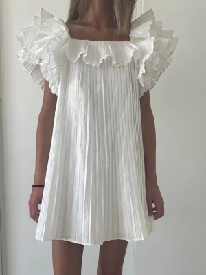 Vit plisserad klänning med volang - Supersöt vit kortklänning med plisserat tyg och stora volanger vid axlarna. Klänningen har en luftig passform och är ärmlös med fyrkantig ringning. Perfekt för dig som gillar en romantisk och trendig look.