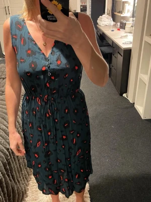 Mörkblå ärmlös klänning med mönster - Säljer en mörkblå ärmlös klänning med v-ringning och knappar framtill. Klänningen har ett coolt leopardmönster i svart och rött samt dragsko i midjan för snygg passform. Perfekt för dig som vill sticka ut lite extra!