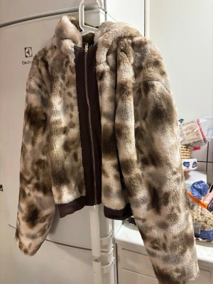 Brun och beige pälsjacka från Cider - Säljer en fluffig pälsjacka från Cider i beige och bruna toner med leopardmönster. Jackan har dragkedja framtill, huva och breda ärmar. Perfekt för dig som vill sticka ut med en cozy och trendig look.