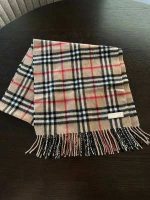 Halsduk från Burberry - Klassisk halsduk från Burberry i 100% kashmir med rutigt mönster i beige, svart, vitt och rött. Halsduken har fransar i ändarna och den ikoniska Burberry-taggen. Perfekt accessoar för att lyfta din outfit. Oanvänd 