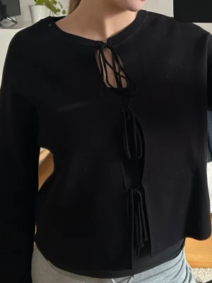 Helt ny! Svart kofta med snörning - Säljer en svart kofta med snygga snörningsdetaljer framtill. Tröjan har lång ärm och är gjord i ett mjukt, stickat material. Perfekt för dig som gillar en unik twist på klassiska plagg.