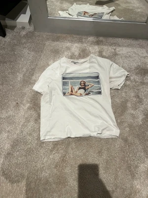 Vit t-shirt med bild  - Vit t-shirt med tryck av en person som solar vid havet på framsidan. Klassisk passform med rund hals och korta ärmar. Perfekt för en chill och avslappnad stil.