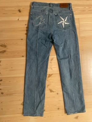 Blå jeans från Denim Genes corteiz  - Säljer ett par blå jeans från Denim Genes i storlek M. Jeansen har raka ben och coola broderade detaljer med stjärnor på bakfickorna. Klassisk femficksmodell med hög midja och tillverkade i bomull. Perfekt för dig som vill sticka ut lite extra.