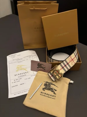 Vändbart burberry bälte med giftboxset - Säljer en Burberry väst med klassiskt rutigt mönster i beige, svart och rött. Västen har en snygg form och är dekorerad med guldiga knappar framtill. Materialet är lyxigt skinn och polyester, perfekt för dig som vill ha en ikonisk look med Burberrys signaturstil.