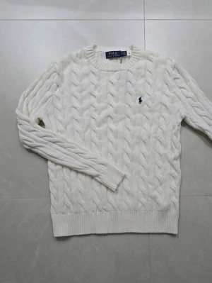 Vit stickad tröja från Polo Ralph Lauren - Säljer en vit stickad tröja från Polo Ralph Lauren med klassiskt kabelmönster och rund hals. Tröjan har långärmade ribbade muddar och en liten broderad logga i marinblått på bröstet. Perfekt för dig som gillar stilren och tidlös design.