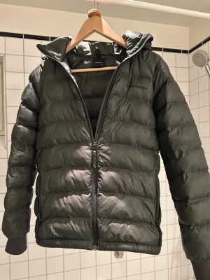 Mörkgrön pufferjacka Peak Performance - Säljer en mörkgrön pufferjacka från Peak Performance med huva och dragkedja framtill. Jackan har quiltad design och ribbade muddar vid ärmslut. Materialet är syntet och jackan är perfekt för kyliga dagar. Finns även en lagad detalj på ryggen.