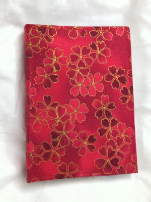 Blomstrande Anteckningsbok - En snygg anteckningsbok med röd tygklädsel och guldmönstrade blommor. Perfekt för dig som vill skriva ner idéer, drömmar eller skisser. Passar både till skolan och fritiden, och är lätt att ta med överallt. Oskrivna sidor ger dig full frihet att skapa din egen berättelse. A6 storlek ca 100 sidor