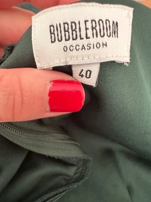Mörkgrön långklänning från Bubbleroom - Snygg mörkgrön långklänning från Bubbleroom Occasion med omlott-detalj vid bysten och slits på sidan. Klänningen är ärmlös och har en figurnära passform. Tillverkad i mjuk polyester med lite stretch för extra komfort. Perfekt för dig som vill ha en elegant look.